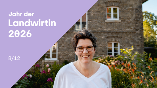 Eine Frau mit kurzen, dunklen Haaren und Brille steht lächelnd in einem Garten vor einem Backsteinhaus. Sie trägt ein weißes Oberteil. Im Hintergrund blühen verschiedene Blumen und Sträucher. Links oben befindet sich ein lilafarbenes Feld mit weißer Schrift: 'Jahr der Landwirtin 2026' und darunter '8/12'. Die Szene wirkt freundlich und einladend.