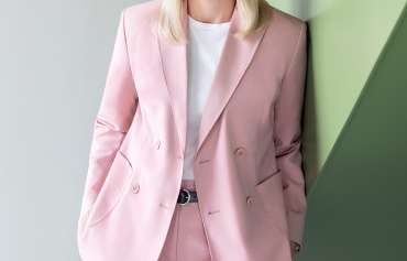 Eine Frau mit glattem, schulterlangem, blondem Haar steht vor einer Wand mit geometrischen Flächen in Hellgrau und Pastellgrün. Sie trägt einen rosafarbenen Hosenanzug mit doppelreihigem Blazer und ein weißes T-Shirt. Ihre Hände sind locker in die Hosentaschen gesteckt. Sie blickt freundlich in die Kamera.