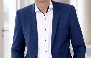 Ein Mann mit kurzem grauem Haar und Brille steht in einem modernen, hellen Innenraum. Er trägt ein dunkelblaues Sakko mit passender Hose, ein weißes Hemd mit dunklen Knöpfen und hat die Hände in den Hosentaschen. Im Hintergrund sind große Fenster und helle Wände zu sehen.