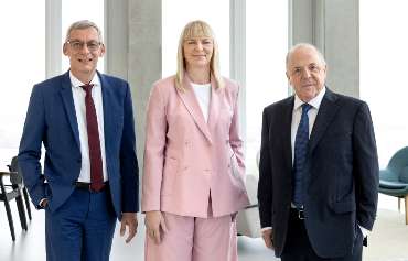 Drei Personen stehen nebeneinander in einem modernen, hellen Raum mit großen Fenstern. Die Person links trägt einen blauen Anzug mit weißem Hemd und roter Krawatte, die Person in der Mitte einen rosafarbenen Hosenanzug mit weißem Oberteil, und die Person rechts einen dunkelblauen Anzug mit weißem Hemd und dunkelblauer Krawatte. Im Hintergrund sind einige Stühle und ein Tisch zu sehen.